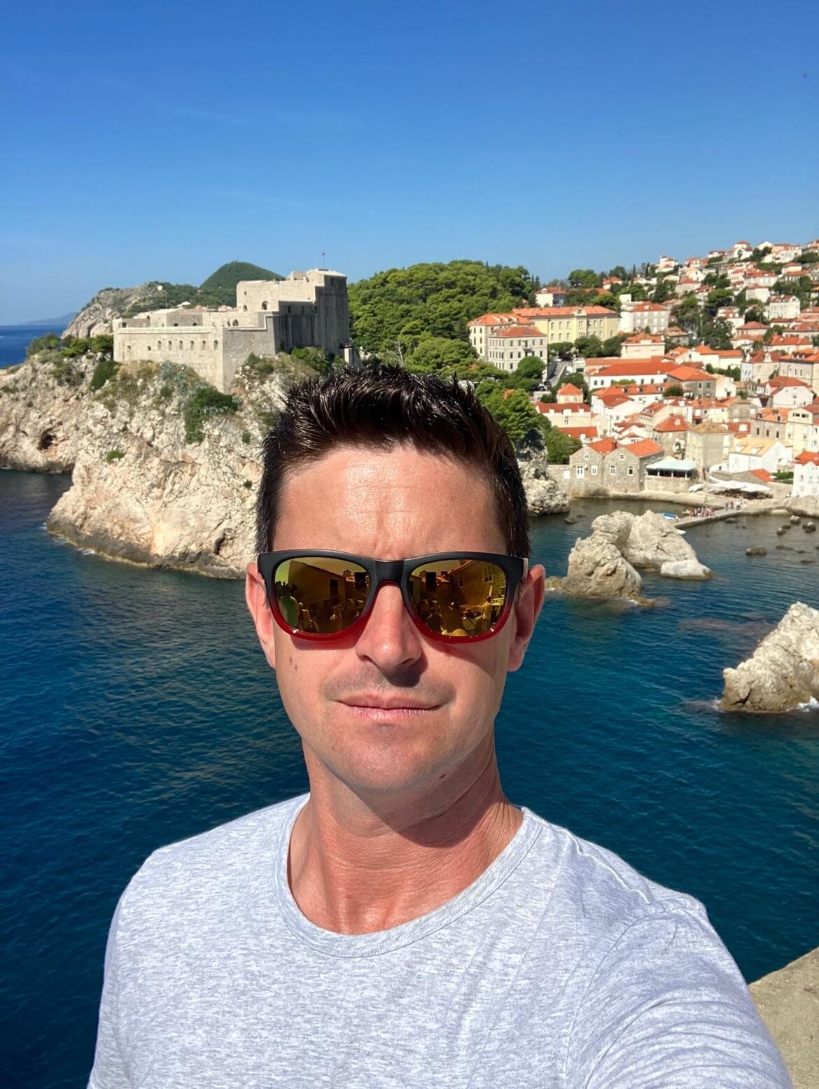 A man wearing sunglasses takes a selfie in front of a coastal town and a stone fortress overlooking the sea.