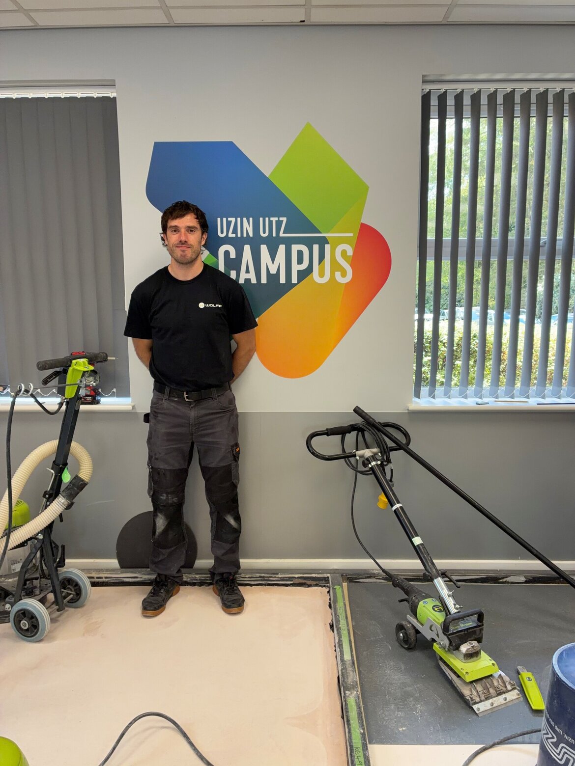 A man stands next to floor sanding equipment in front of a colorful UZIN UTZ CAMPUS logo on the wall, with large windows and blinds in the background.