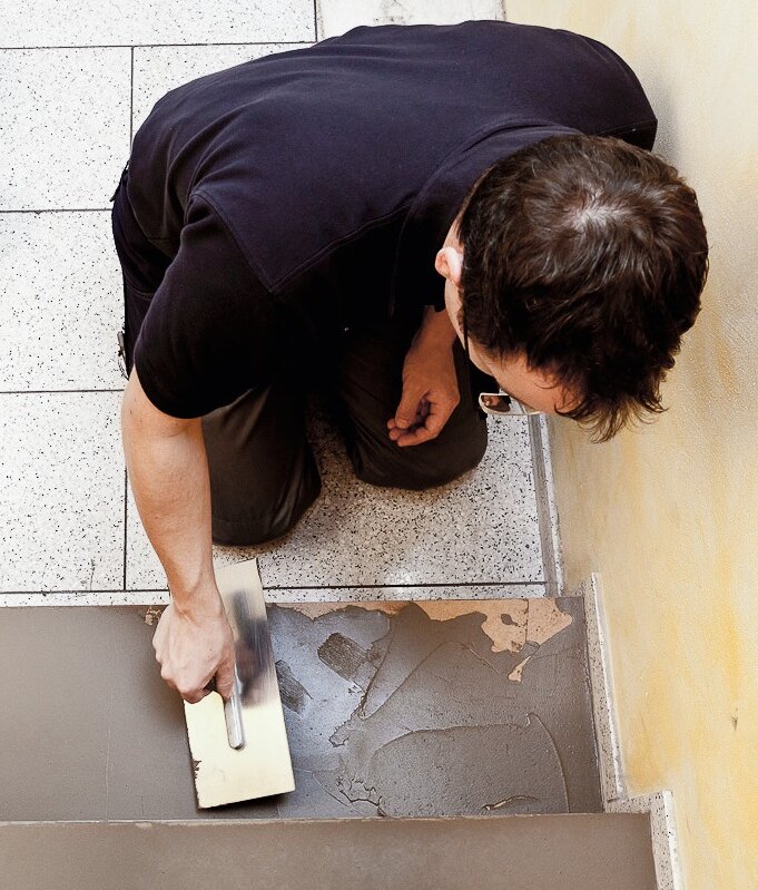 A person kneels on tiled stairs, using a trowel to spread UZIN NC 888 F Fibre Finish on a stair tread next to a yellow wall. A person kneels on tiled stairs, using a trowel to spread UZIN NC 888 F Fibre Finish on a stair tread next to a yellow wall.